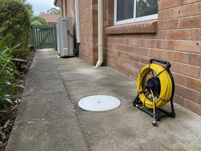 Capped drain inspection opening beside a yellow camera reel on a concrete side path by a brick house in Cherrybrook