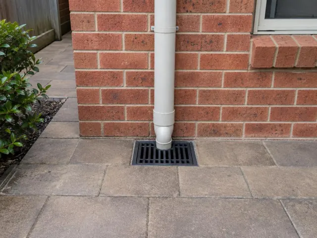 Brick house wall with a seated stormwater grate and dry paving beside a downpipe in Winston Hills